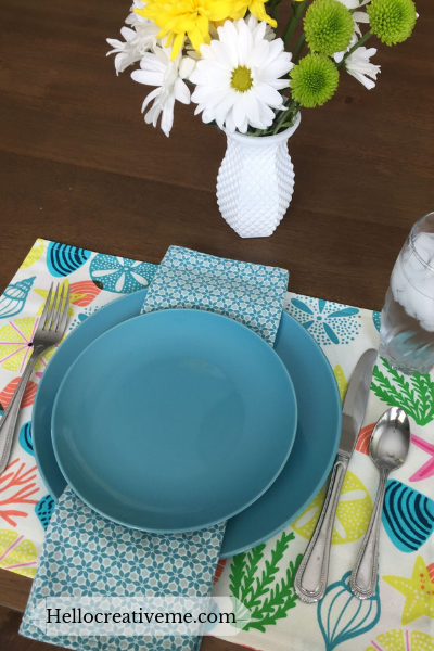 Table set with teal blue dishes on multicolored DIY placemat and vase of flowers