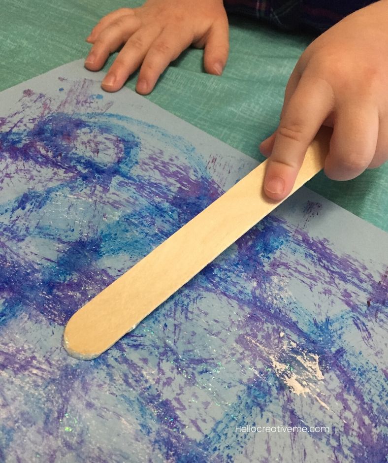 boy spreading white glitter glue on paper using popsicle stick