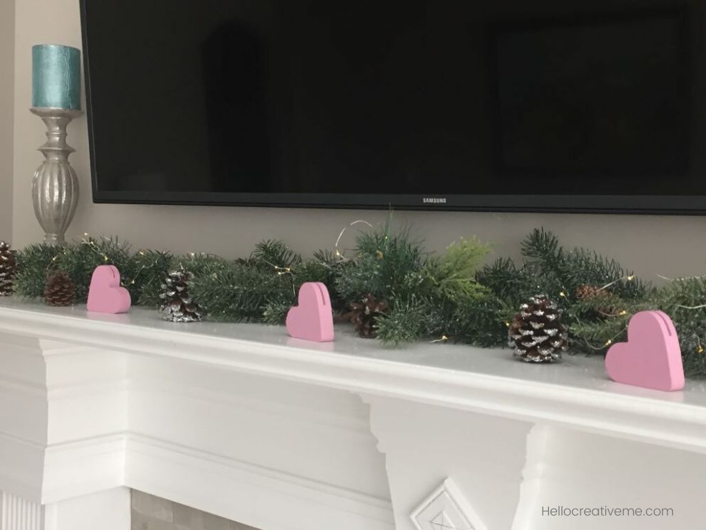 Three pink wooden hearts with green garland on fireplace mantle.