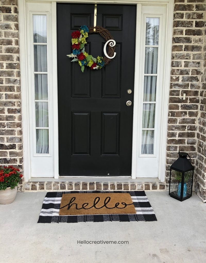 A refreshed spring front porch with colorful wreath on black door and hello mat layered with black and white stripe rug.