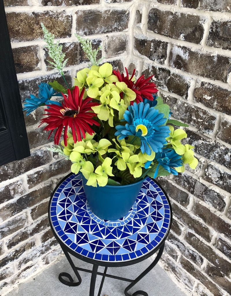 Faux burgundy, green, and blue flowers in a teal pot on a plant stand.